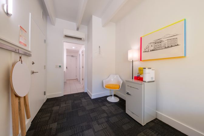 Bright modern hotel entryway with white mid-century chair with yellow cushion, retro-style mini fridge and coffee maker, floor lamp, framed architectural wall art with colorful trim, air conditioner above doorway, and dark patterned carpet.