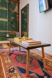 Mid-century wooden coffee table with tapered legs holding a yellow tray, small potted cactus and book on a colorful Persian-style rug in an industrial living room with distressed green plaster wall, mirror and hardwood floors.