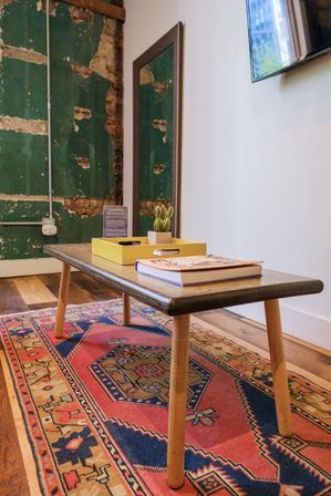 Mid-century wooden coffee table with tapered legs holding a yellow tray, small potted cactus and book on a colorful Persian-style rug in an industrial living room with distressed green plaster wall, mirror and hardwood floors.