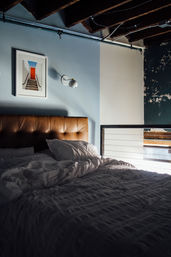 Cozy urban loft bedroom with tufted brown leather headboard, rumpled white bedding, blue accent wall, modern wall sconce and exposed industrial ceiling