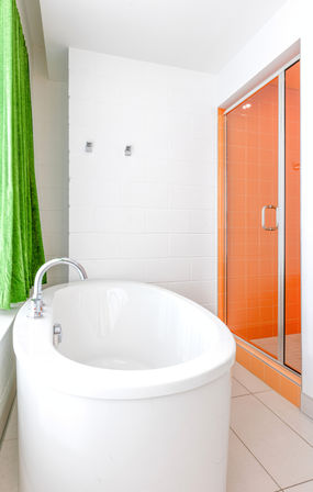 Bright modern bathroom with white freestanding oval bathtub and chrome faucet, vibrant green curtain, and orange-tiled glass shower enclosure