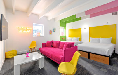 Bright modern hotel suite with a hot-pink sofa, two white beds with bold yellow headboards, yellow chairs, white coffee table, neon wall sign, green accent wall, exposed beams and playful colorful decor