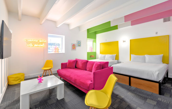 Bright modern hotel suite with a hot-pink sofa, two white beds with bold yellow headboards, yellow chairs, white coffee table, neon wall sign, green accent wall, exposed beams and playful colorful decor