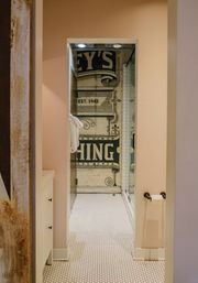 Cozy boutique-style bathroom corridor with white hexagon tile floor, pale pink walls, glass shower featuring a vintage green graphic mural reading “EST. 1942,” folded white towels on a hook and a toilet paper holder.