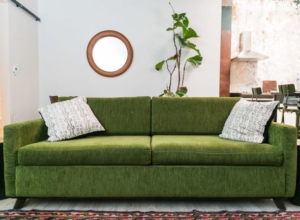 Lush green mid-century modern sofa with patterned throw pillows in a bright living room, round wall mirror above, tall potted plant beside it and an open-plan kitchen visible in the background