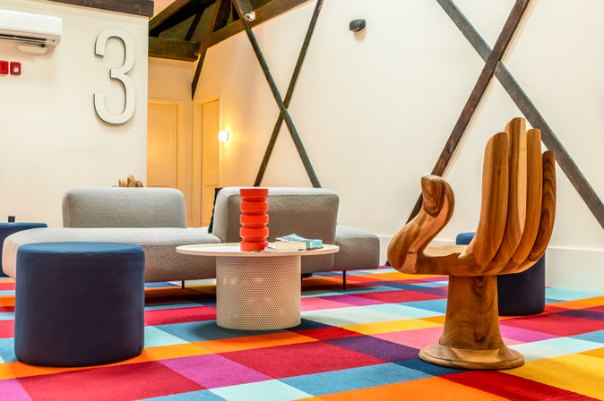 Bright modern lounge area with multicolored checkerboard carpet, low gray sofas and navy ottomans, round white coffee table with a red stacked vase and books, sculptural wooden hand chair, exposed dark beams and a large number 3 on the white wall.