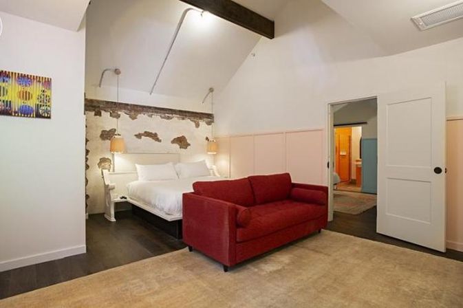 Bright loft-style bedroom with vaulted ceiling, king bed against a distressed plaster accent wall, red loveseat on a beige rug, dark hardwood floors and open doorway.