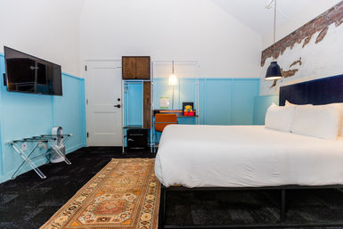 Bright boutique hotel room with king bed and navy headboard, blue wainscoting, exposed brick accent, wall-mounted TV, small desk with orange chair and hanging pendant, patterned runner rug.