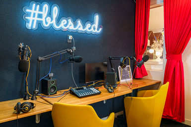 Podcast studio setup with neon "#blessed" sign on a dark soundproof wall, dual boom microphones, mixing console, monitor, yellow chairs and red curtains.