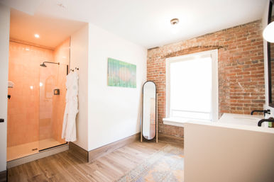 Bright loft-style bathroom with exposed red brick wall and large window, glass walk-in shower with pale pink tiles, white robe on a hook, oval floor mirror, black fixtures and wood-look tile floor.