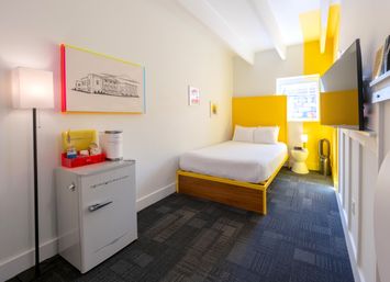 Bright modern boutique hotel room with yellow accent headboard and trim, white bed, wall-mounted TV, retro mini-fridge and floor lamp in a compact urban layout