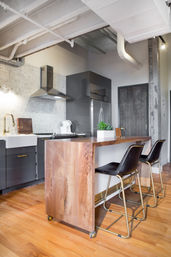 Modern industrial loft kitchen with wooden island on casters, two black leather bar stools with gold legs, gray cabinets, stainless refrigerator and range hood, white farmhouse sink, hex tile backsplash, hardwood floors and a small succulent centerpiece.