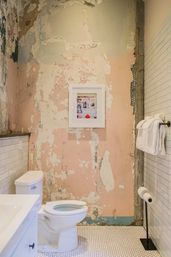 Bathroom interior with white toilet and sink, peeling pink plaster wall with framed art, white subway tile wainscoting, rolled towels on a bar and white hex floor tiles — rustic, distressed-chic style.