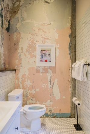 Bathroom interior with white toilet and sink, peeling pink plaster wall with framed art, white subway tile wainscoting, rolled towels on a bar and white hex floor tiles — rustic, distressed-chic style.