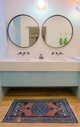 Modern bathroom double vanity with two round mirrors, matte black wall-mounted faucets, pale blue tiles, white countertop, and a vibrant patterned rug on a wood floor.