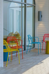 Bright yellow, red, and blue metal patio chairs on a sunlit tiled terrace outside a modern white building with large glass sliding doors