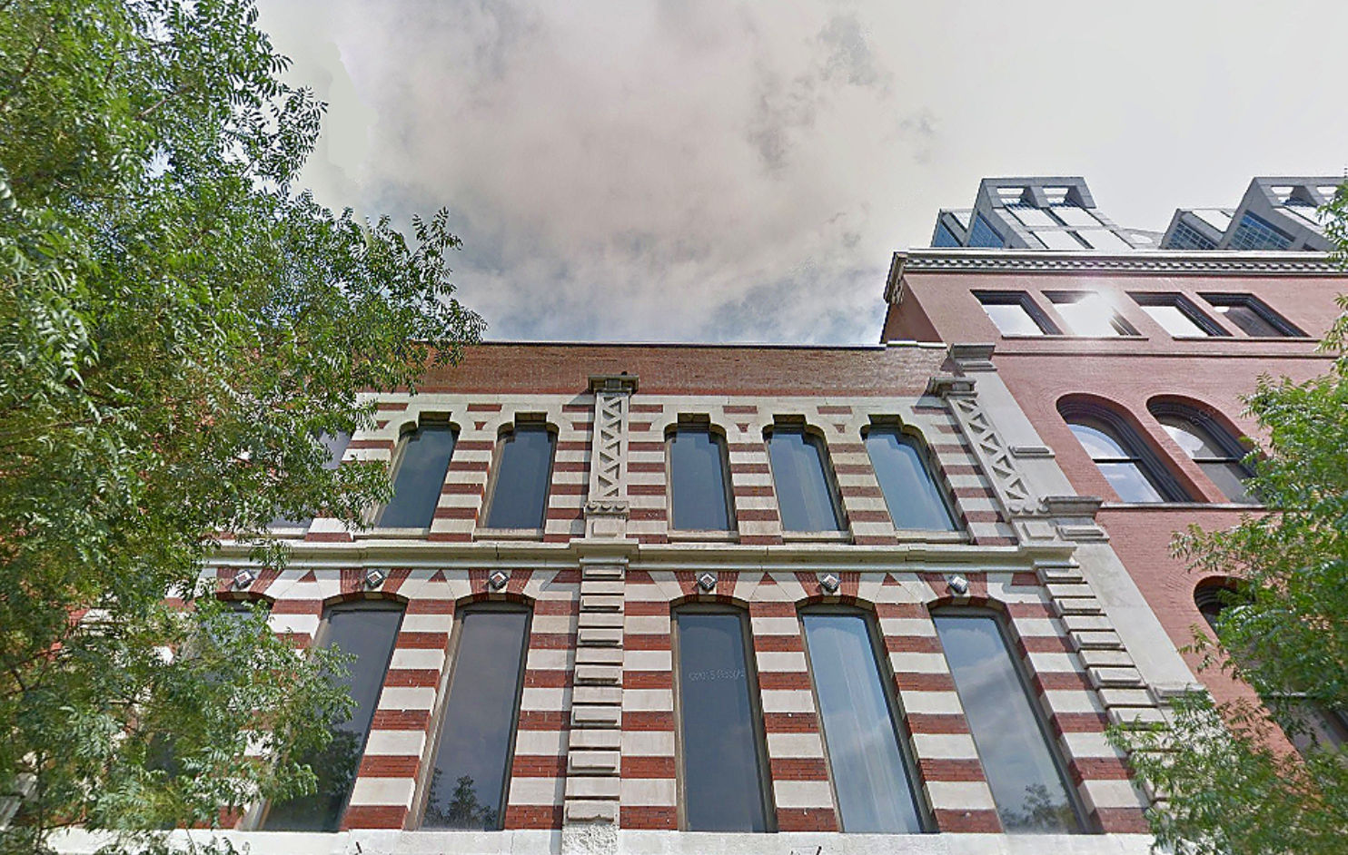 Historic downtown red-and-white striped brick building with tall arched windows, decorative stone trim, leafy trees in front and modern rooftop additions peeking above