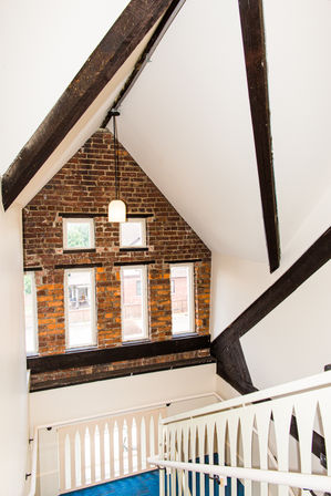 Lofty stairwell in a converted historic brick townhouse with exposed dark wooden beams, a brick gable wall of tall narrow windows, a hanging pendant light, white arched railing and blue carpeted stairs.