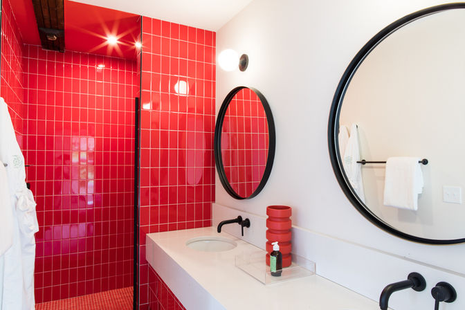 Modern bathroom with glossy red tiled walk-in shower, white countertop vanity, two round black-framed mirrors, matte-black wall-mounted faucets, stacked red vase and soap dispenser, white towels and robe.