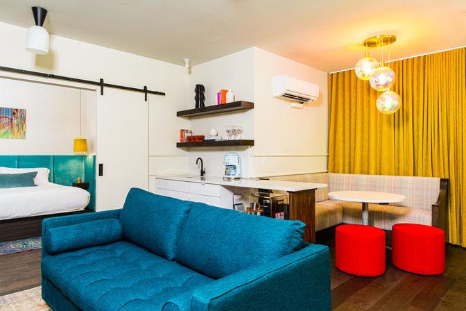 Bright, modern studio interior with teal sofa, bed behind a sliding barn door, compact kitchenette, plaid banquette, round table with red stools, mustard-yellow curtains and globe pendant lights.