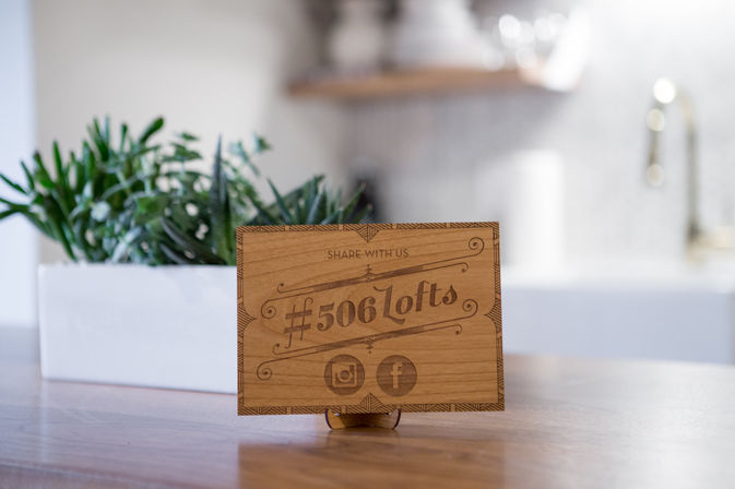 Engraved wooden tabletop sign with a hashtag and social media icons on a small stand, sitting on a wood countertop next to a white planter of succulents with a blurred modern kitchen sink in the background.