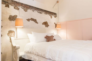 Cozy bedroom interior with a large white bed and fluffy duvet, exposed distressed plaster and brick accent wall, soft pink wall panels, two warm hanging pendant lamps, and a small rust-colored throw pillow.