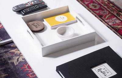 White modern coffee table with a recessed tray holding a yellow notebook, round coasters and a small marble bowl; TV remotes and a black photo album sit nearby on colorful patterned rugs