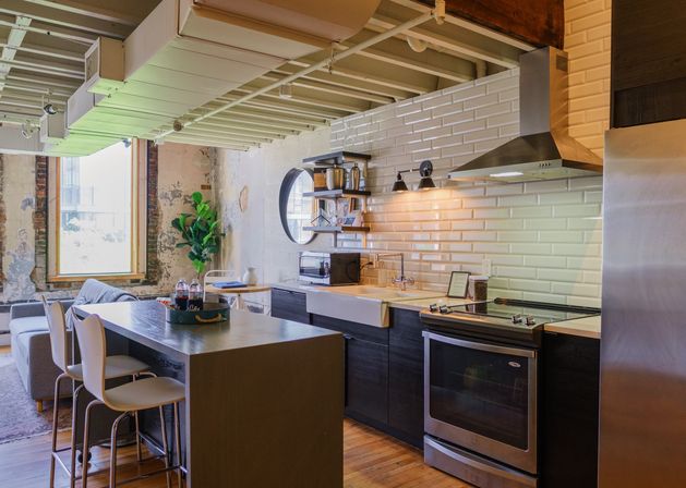 Open-plan industrial loft kitchen with white subway tile backsplash, stainless steel range and hood, farmhouse sink, dark wood cabinets, breakfast island with two white bar stools, hardwood floors, large window and exposed distressed brick walls for a cozy urban vibe.