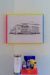 Framed black-and-white architectural sketch in a rainbow acrylic frame above a hotel guest welcome station with a single-serve coffee maker, bottled water, snacks and a guest guide.