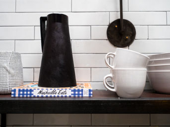 Black ceramic pitcher atop a "Nashville Eats" cookbook next to stacked white mugs and bowls on a kitchen shelf with white subway tile backsplash