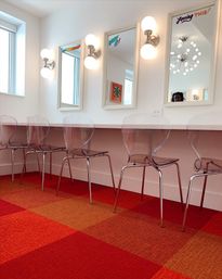 Bright salon-style vanity row with clear acrylic chairs, white-framed mirrors with globe wall sconces and a red-orange checkered carpet