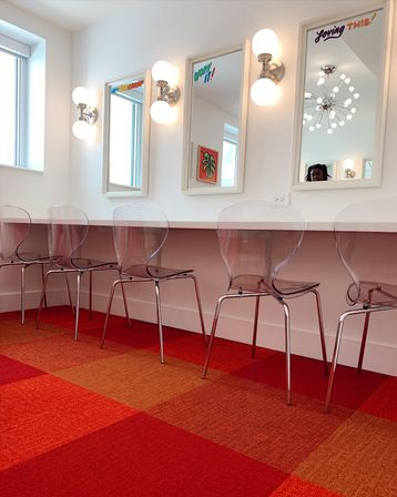Bright salon-style vanity row with clear acrylic chairs, white-framed mirrors with globe wall sconces and a red-orange checkered carpet