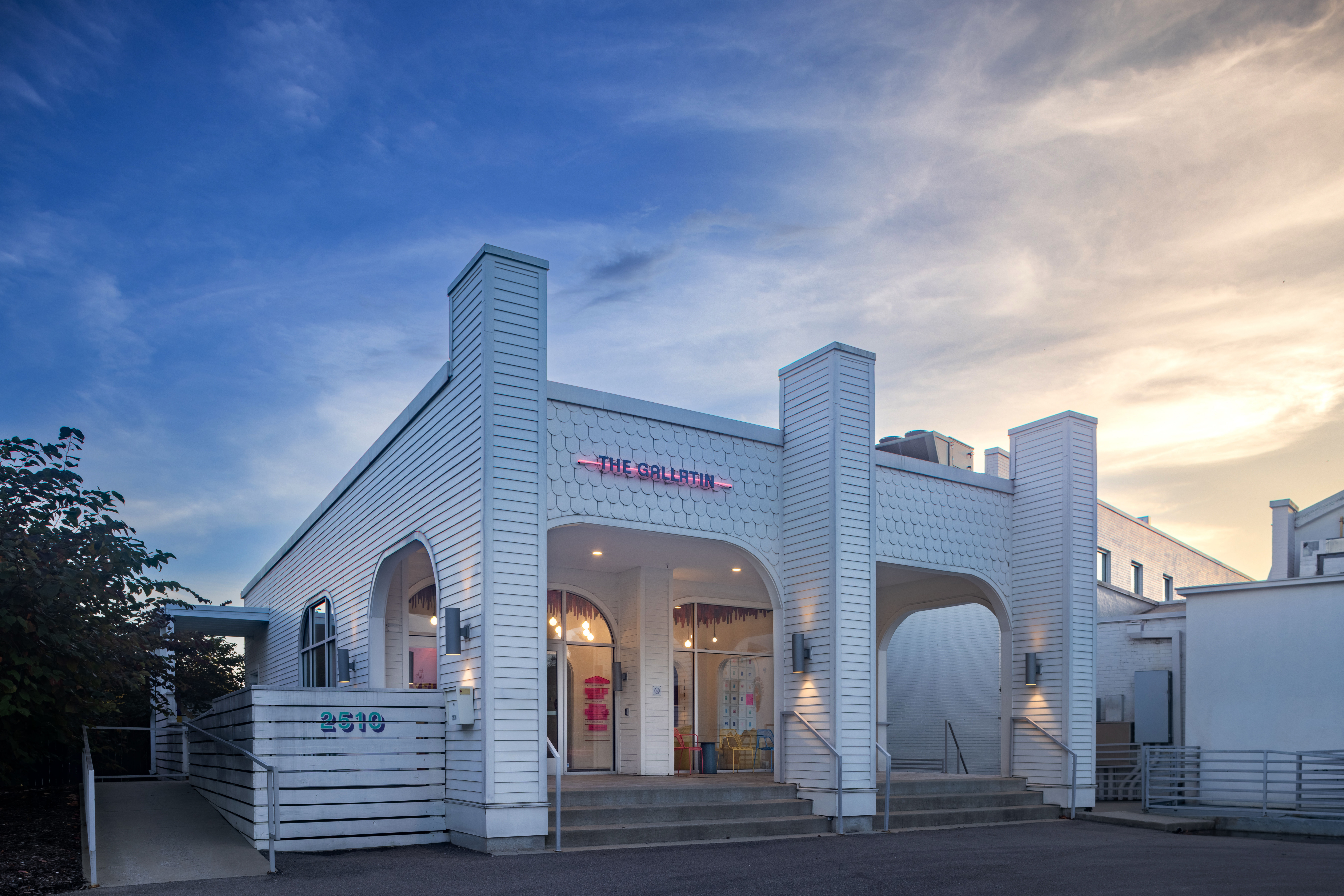 Charming white coastal-style storefront with arched entryways, steps and wheelchair ramp, pink neon sign above the entrance and pastel-lit seating inside under a blue and golden sunset sky