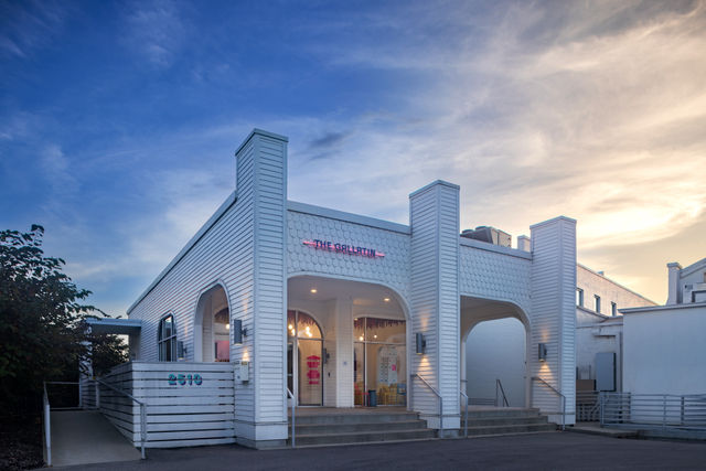 Charming white coastal-style storefront with arched entryways, steps and wheelchair ramp, pink neon sign above the entrance and pastel-lit seating inside under a blue and golden sunset sky