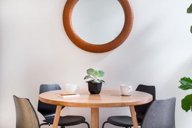 Bright, cozy dining nook with a round wooden table, four black chairs, a small potted succulent at the center, two white coffee mugs and a round leather-framed wall mirror.