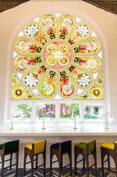 Vibrant stained-glass rose window flooding a historic arched interior with sunlight over a white counter lined with colorful bar stools and brass reading lamps, street view visible through lower panes.