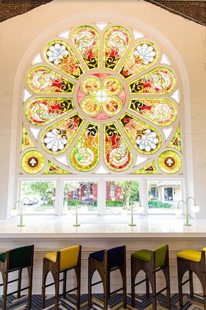 Vibrant stained-glass rose window flooding a historic arched interior with sunlight over a white counter lined with colorful bar stools and brass reading lamps, street view visible through lower panes.