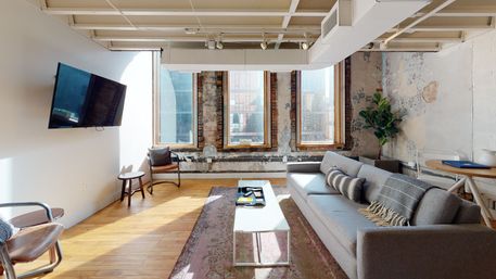 Sunlit industrial loft living room with exposed brick window frames, hardwood floors, gray sofa with patterned pillows, white coffee table on a faded rug, wall-mounted TV and potted plant in an urban apartment interior