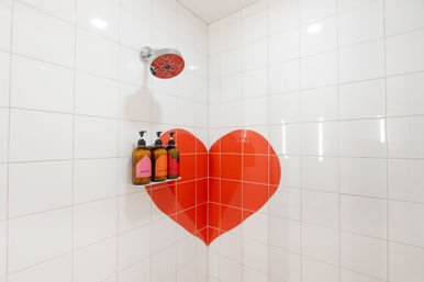White tiled corner shower with a large red heart tile accent, chrome showerhead, and amber pump bottles of shampoo, conditioner and body wash on a small wall shelf