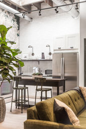 Modern loft kitchen with white subway tile, stainless steel refrigerator, dark wood island with metal bar stools, large fiddle-leaf plant and green sofa in foreground.