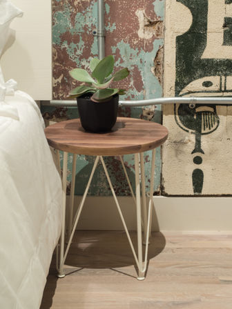 Round wooden bedside table with a black-potted succulent by a white bed, set against a distressed industrial wall with exposed conduit and painted mural on a hardwood floor