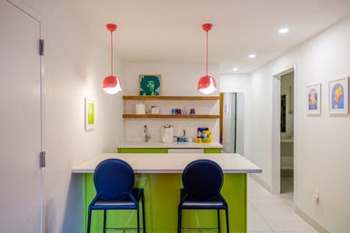 Bright modern apartment kitchenette with lime-green island and white quartz countertop, two navy bar stools, playful red pendant lights, open wood shelves with sink and coffee maker, and colorful framed art