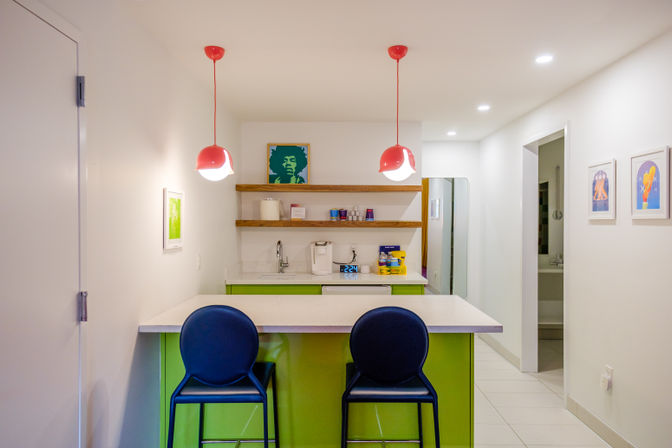 Bright modern apartment kitchenette with lime-green island and white quartz countertop, two navy bar stools, playful red pendant lights, open wood shelves with sink and coffee maker, and colorful framed art