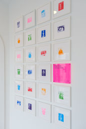 Minimalist white gallery wall in a modern interior showing a grid of small white-framed colorful photo prints and a neon-pink acrylic plaque that reads 'Stay here, change lives'.