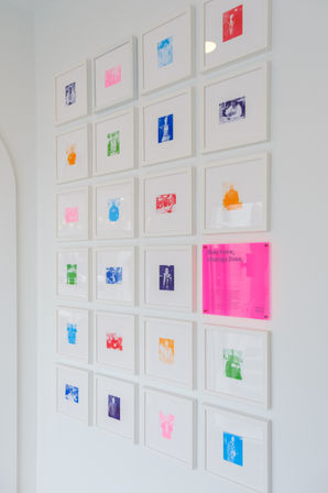 Minimalist white gallery wall in a modern interior showing a grid of small white-framed colorful photo prints and a neon-pink acrylic plaque that reads 'Stay here, change lives'.