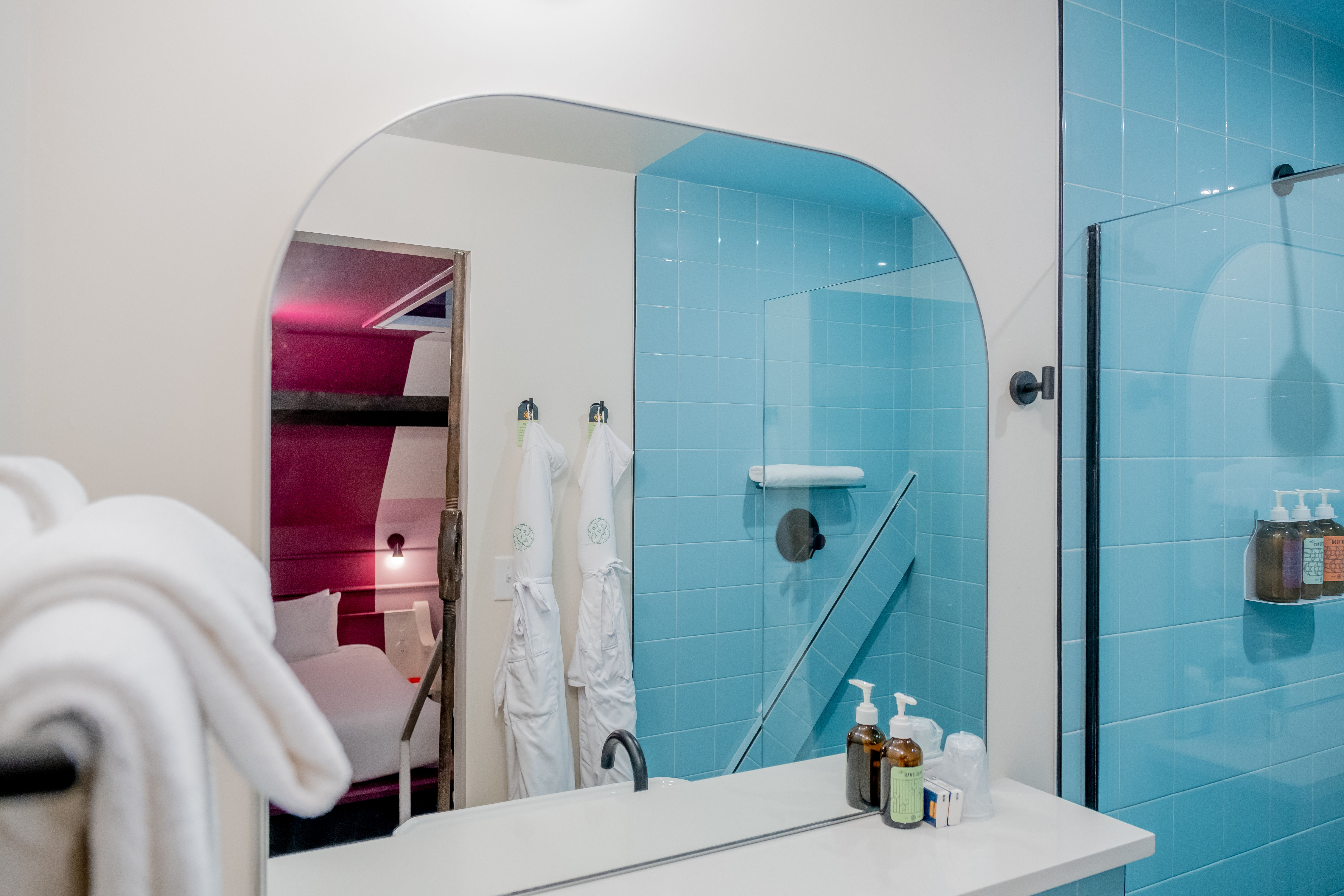 Modern bathroom vanity mirror reflecting aqua-blue tiled glass shower, white counter with soap dispensers and towels, hanging white robes and a pink-lit bedroom glimpse