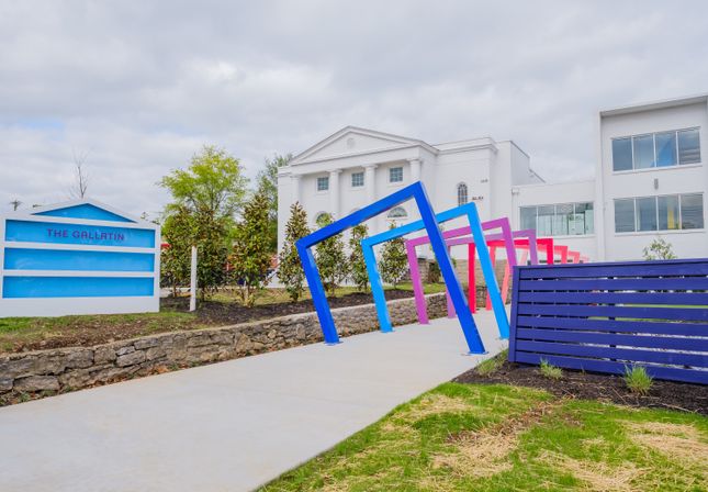 Vibrant blue, pink, and purple tilted archway over a sidewalk leading to a white columned classical-style building, stone landscaping, and modern outdoor seating on an overcast day.