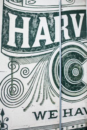 Vintage-style green mural on a white brick wall featuring large decorative letters (partial "HARV"), ornate swirls and concentric rings, with a vertical metal conduit running down the center.
