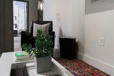 Cozy modern living room corner with a potted succulent on a white coffee table, stacked books and a remote, black armchair with a textured pillow, hanging pendant light and a colorful patterned rug.