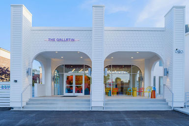White retro-modern inn entrance with twin arched porches, wide concrete steps leading to glass double doors, colorful metal chairs on the covered patio and pendant lights under a clear blue sky.